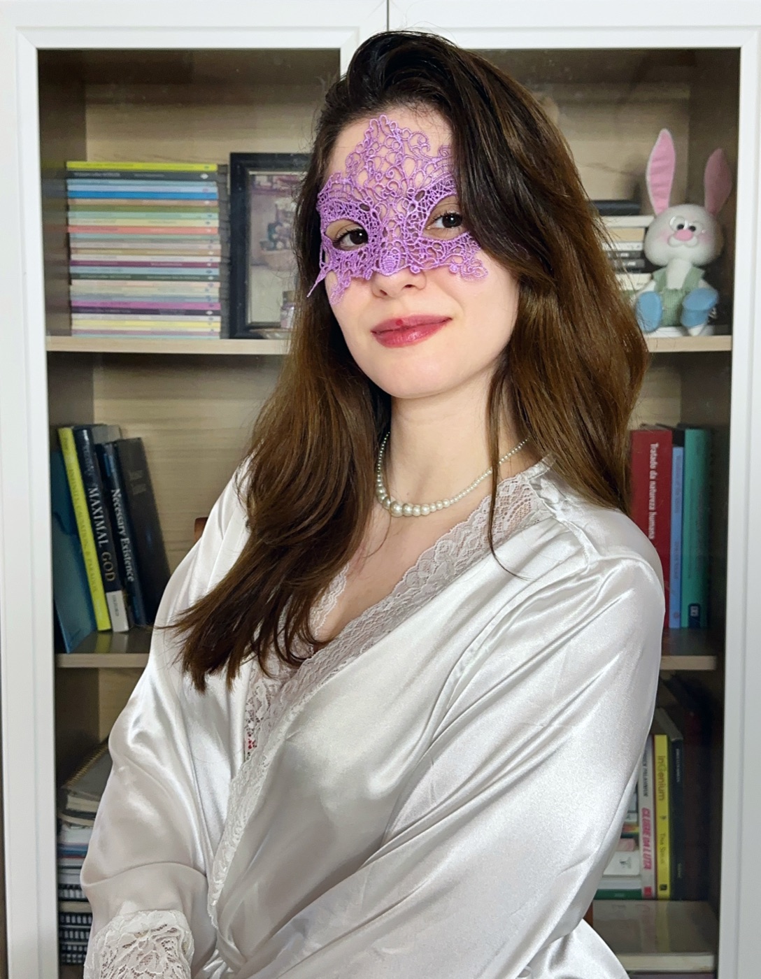 Liza, de máscara de renda lilás e robe de cetim, diante da sua estante de livros